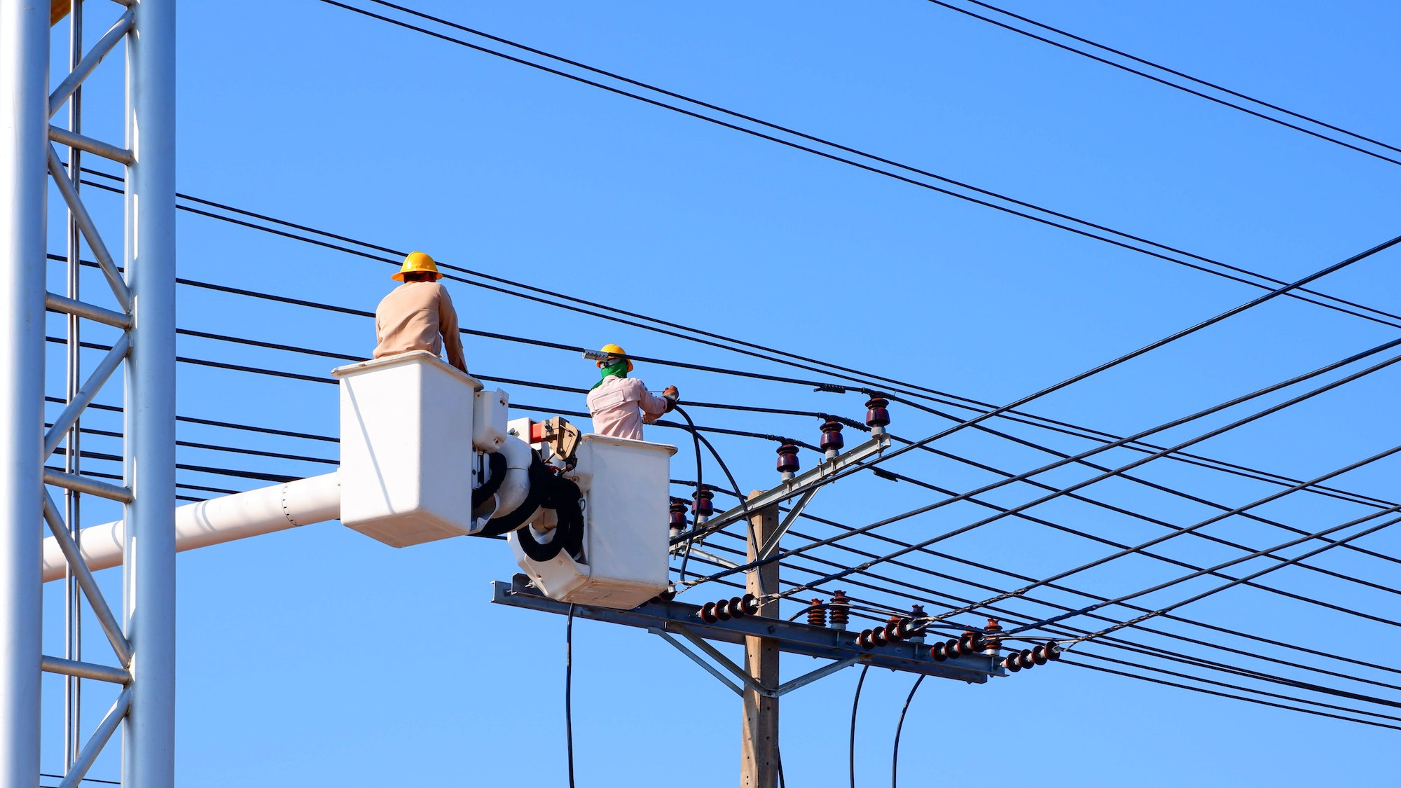Two electricians on bucket boom truck installing electrical system on power pole against blue sky